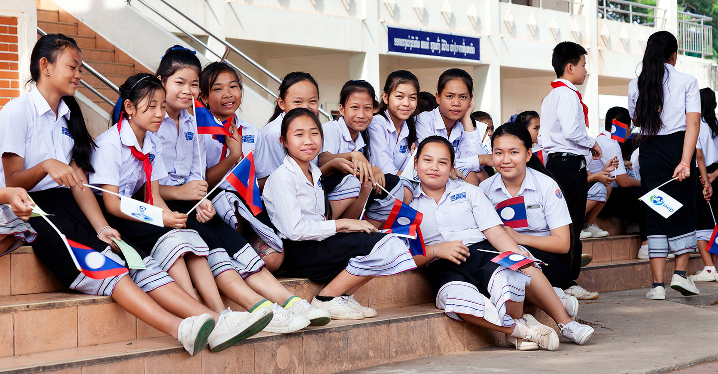 School girls smile for a picture while holding the Laotian flag with the Gavi logo on the back