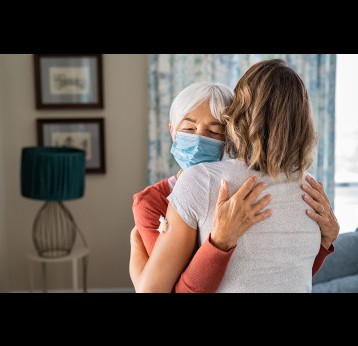 Senior woman wearing face mask hugging immunized daughter after covid-19 vaccination shot.