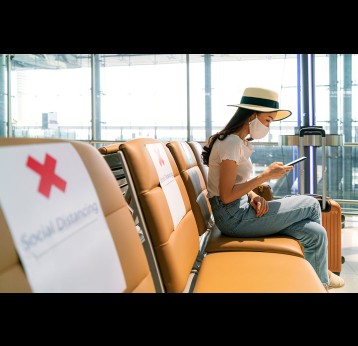 Person wearing mask using mobile phone searching airline flight status and sit social distancing chair in airport