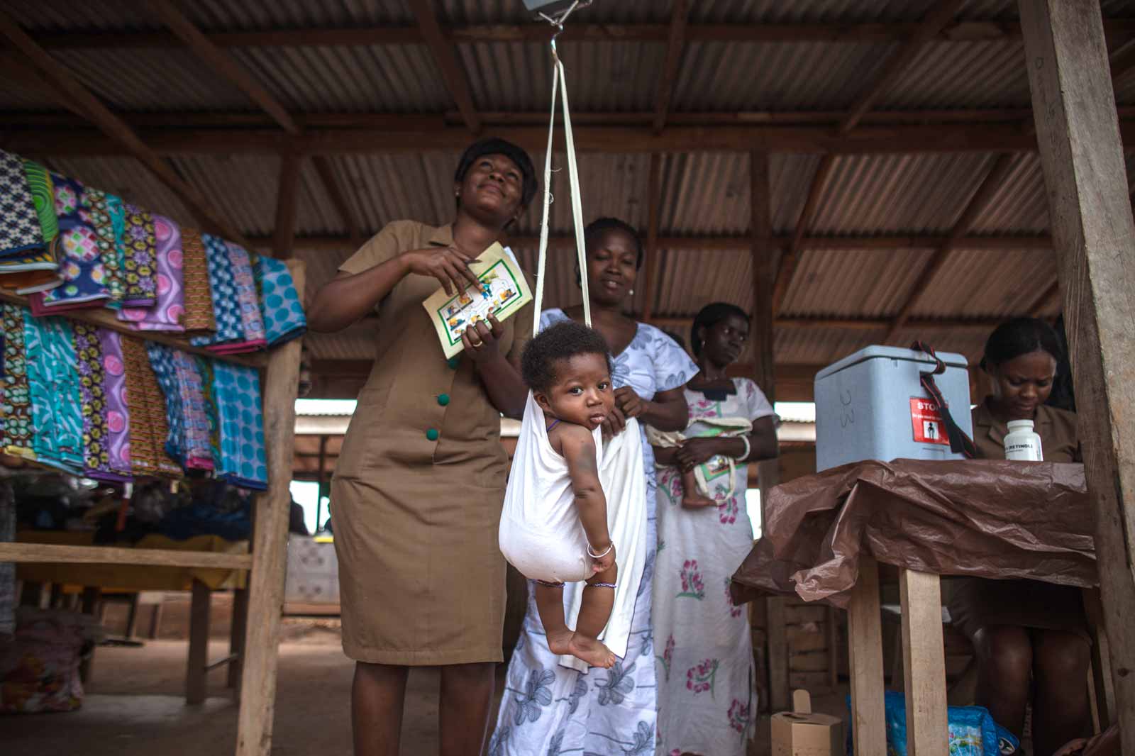   Au dispensaire installé sur un étal vide de marché, l’agent de santé communautaire Mavis Adjetey pèse les bébés avant d’administrer les vaccins. En 2012, le Ghana est devenu le premier pays d’Afrique à introduire simultanément des vaccins contre la pneumococcie et le rotavirus, ciblant ainsi les premières causes de mortalité dans le monde : la pneumonie et la diarrhée.