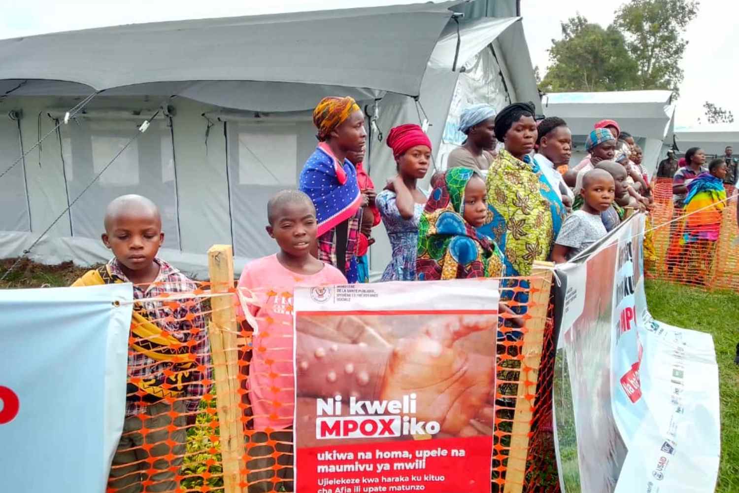 In the DRC, families wait for mpox vaccination at a health centre. A poster in Swahili informs of the symptoms and encourages medical consultation. Credit: Ricky Ombeni